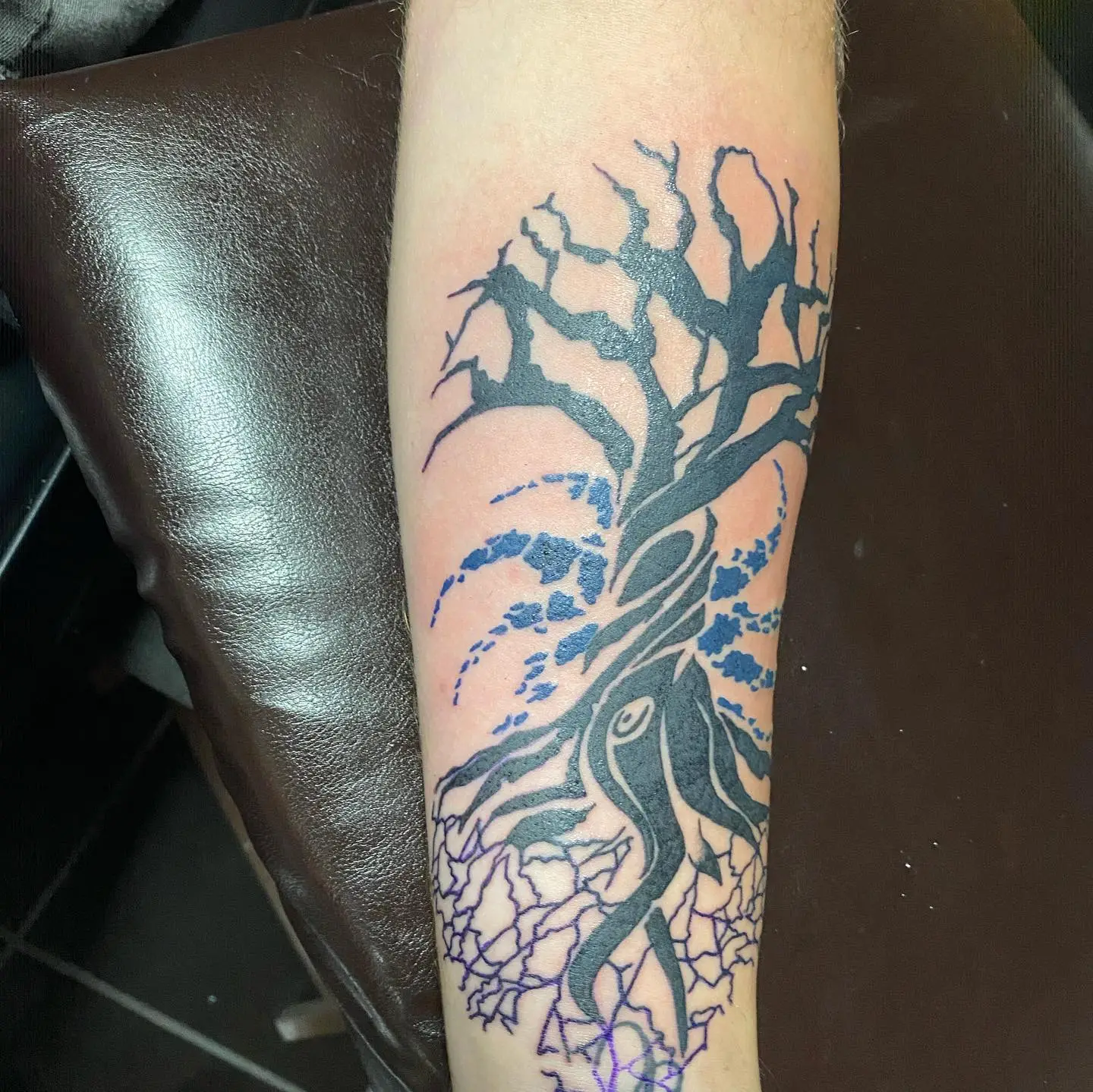 Abstract tattoo of a twisted tree with both roots and branches showing, and a blue swirling design behind the tree.