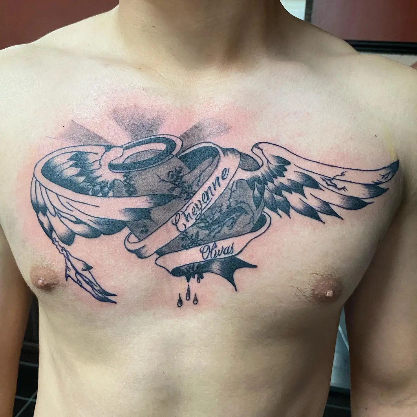 Chest tattoo of a heart with a halo, angel wings and a banner wrapped around it.